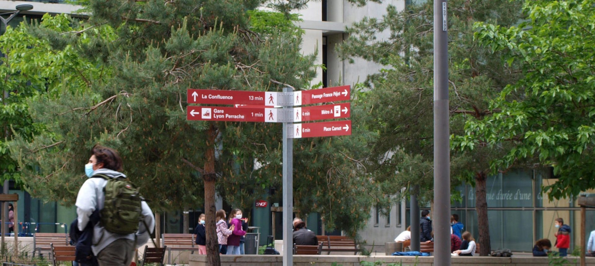 Panneau directionnel rouge du pôle d’échanges Lyon Perrache au milieu d’arbres et de passants sur une place